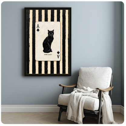 Framed artwork of a black cat inside an ace playing card beside a pale reading chair against a gray wall.