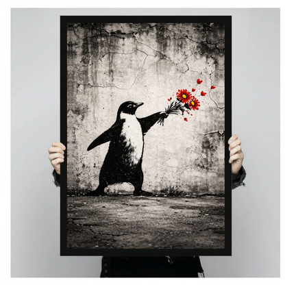 A person holds black framed penguin offering flowers canvas against a light neutral background.