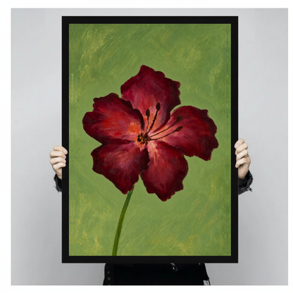 A person holds black framed burgundy bloom on olive canvas against a light neutral background.