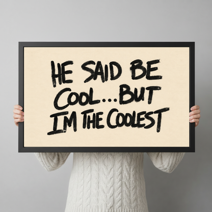 A person wearing a cream sweater holds black framed he said be cool quote canvas in both hands.