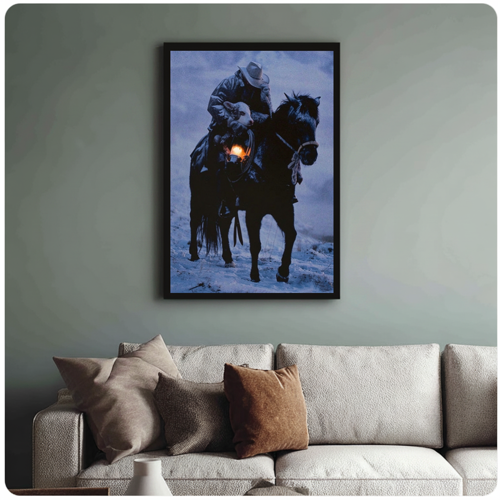 Framed cowboy carrying lamb canvas above a neutral sofa with cushions against a muted wall.