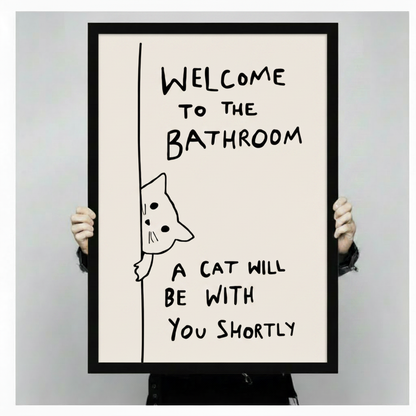 Two hands holding a large black-framed canvas print of a hand-drawn peeking cat with funny bathroom welcome quote, showing print size and frame quality