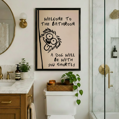 Quirky dog bathroom wall art framed in black hung above toilet in a warm luxury bathroom with gold fixtures marble countertop round mirror and glass shower enclosure