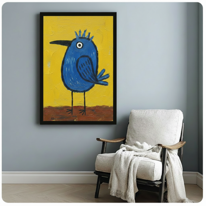 Black framed canvas of a folk-art style round blue bird with a spiky crown and sharp black beak on a bright yellow textured background, mounted on a blue-grey wall beside a white linen armchair