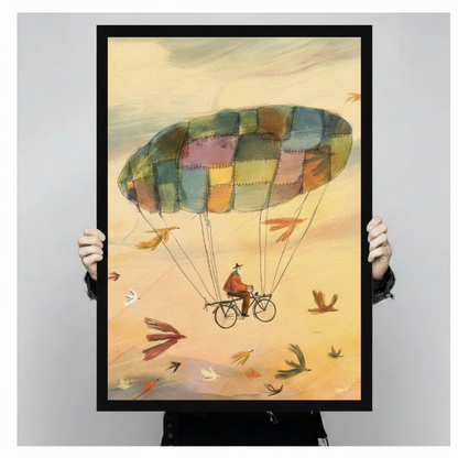 Person holding a large black framed whimsical watercolor illustration canvas art print showing print size scale with an old man cycling through a warm golden sky suspended from a colorful patchwork parachute with flying birds all around
