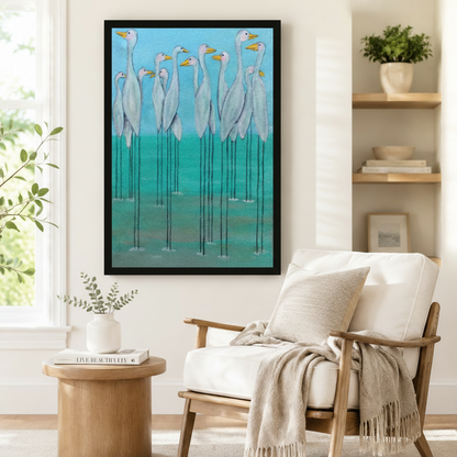 Black framed whimsical white egret flock canvas art showing a gathering of elegant white egrets with bright yellow beaks and spindly black legs standing in shallow turquoise water against a vivid sky blue and teal painterly background, displayed beside a white linen armchair in a cozy reading nook