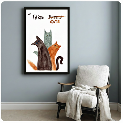 Black framed whimsical The Three Cats watercolor canvas art featuring a trio of cats in dark brown teal-gray and rust orange with wide quirky eyes grouped closely together with expressive hand-lettered The Three Fates crossed out Cats title in loose watercolor style on a white background, hung on a blue-gray wall beside a white linen folding chair