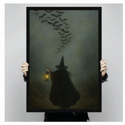 Close-up product shot of a black framed gothic witch and bats wall art print held by two hands, showcasing the eerie foggy lantern scene in detail