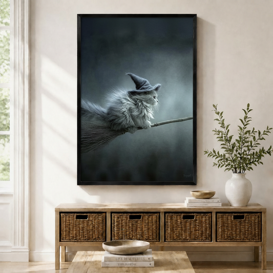 Framed gothic wall art print of a fluffy gray cat wearing a witch hat riding a broomstick, displayed above a wicker console table in a bright entryway