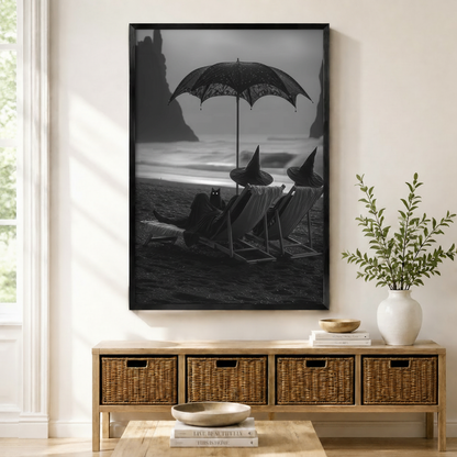 Framed black and white gothic wall art print of witches relaxing on a dark beach with a black cat, displayed above a wicker sideboard in a neutral entryway