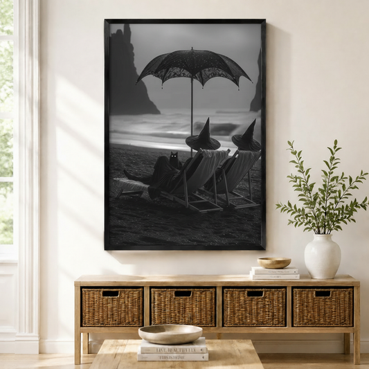 Framed black and white gothic wall art print of witches relaxing on a dark beach with a black cat, displayed above a wicker sideboard in a neutral entryway