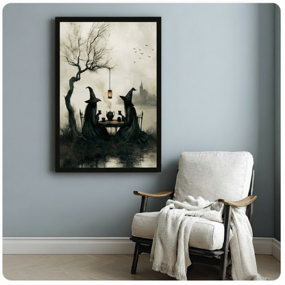 Black framed gothic witches tea party illustration with misty lake and haunted castle background hanging on light blue-gray wall beside a cozy white linen armchair