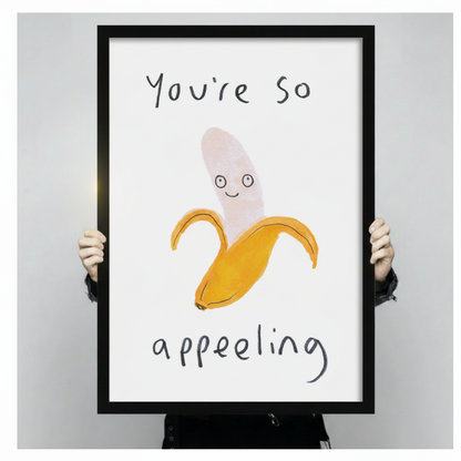 Hand-held black framed canvas print of whimsical smiling banana with "You're So Appealing" hand-lettered pun on white background